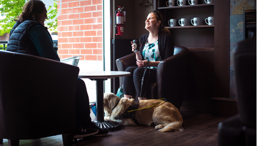 Larissa is sitting in a chair across from another person in a coffee shop by the window with her guide dog Piper laying on the floor at her feet.