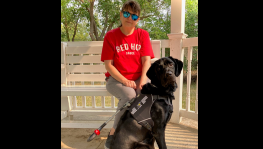 Jodi is sitting on a bench in a gazebo outdoors. Shadow, a black Labrador-golden-retriever mix, is sitting on the ground in front of her.