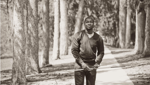 A black and white photograph of Ben walking in a forest. He holds his white cane up in the air near his waist.