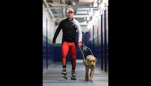 Kevin wearing ice skates and sports clothing walking down a corridor with his guide dog.