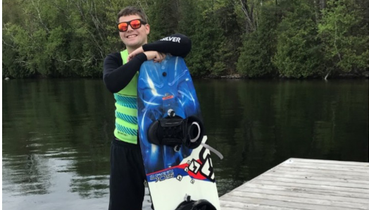 Josh est debout sur le bord du quai, vêtu d'une chemise et d'un short noirs, avec un gilet de sauvetage vert. Un ensemble de skis nautiques bleus et jaunes est posé sur le quai, et il est appuyé sur une planche nautique. Il sourit et porte des lunettes de soleil.