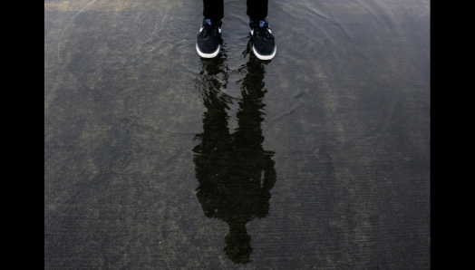 Un jeune homme se tient debout et regarde au-dessus d'une flaque d'eau. Ses chaussures de course apparaissent en haut de l'image. Dans la flaque d'eau apparaît un reflet délicat de sa silhouette.
