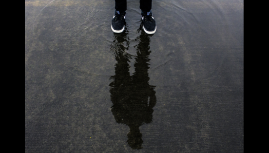 A young man stands peering over a puddle. Their running shoes appear at the top of the image. In the puddle is a soft reflection of their silhouette.