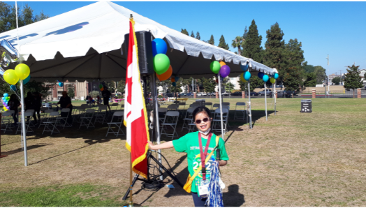 Janna stands beside a Canadian flag outside a large outdoor event tent. She smiles and holds a pom-pom in her left hand.