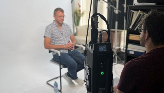 A behind-the-scenes photo of a man sitting in a chair in front of a film camera. A large production light is in the forefront of the photo, next to the Director, whose back is turned to the camera