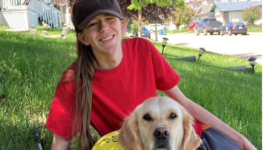 Zach est allongé sur l'herbe avec son chien Elsie, un golden retriever qui porte un gilet jaune d'INCA. La canne blanche de Zach est posée sur l'herbe près de lui.