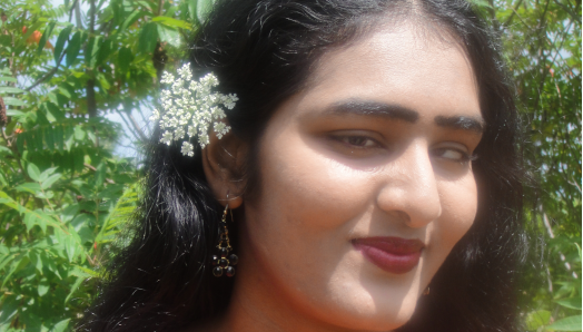  Headshot of Kamini, who is smiling and wearing a red v-neck top and a white flower in her hair.