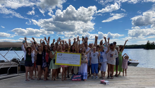 Un groupe d'invités debout sur un quai au bord de l'eau et salue avec enthousiasme, en montrant leur don d’un chèque géant de 1 000 $. 