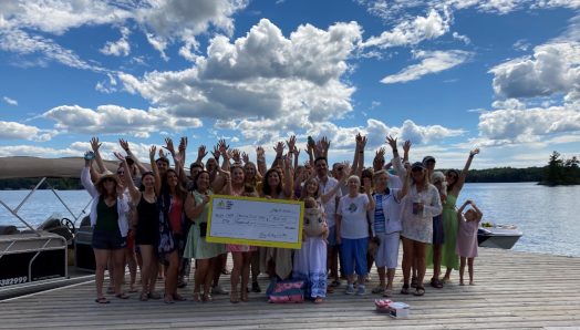 A group of guests stand on top of a waterfront dock and enthusiastically wave at the camera, showcasing their giant-cheque donation of $1,000 