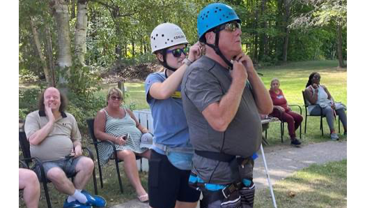 Un membre du personnel aide à fixer le casque d'un invité au pied de la tour d'escalade du camp. 