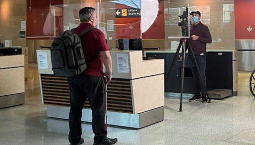 Duane Morgan, CNIB’s Vice President of Atlantic Canada, stands in front of an airport kiosk with a cameraman filming in the background.