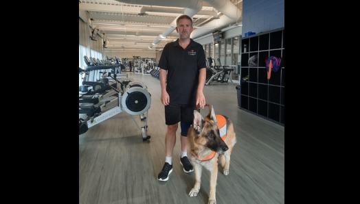 Bob et son chien-guide Berger allemand à l’intérieur d’un gymnase. La salle de gym est remplie d’équipement d’entraînement.