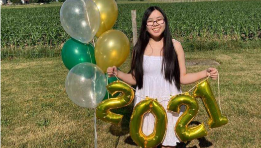 Stormy stands in front of a rural corn field on a sunny summer day. She wears a sundress and is holding a bouquet of graduation-themed balloons