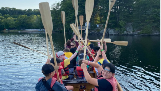  Du canotage au Centre Lake Joe! Des enfants et des jeunes sont assis dans le canoë Voyageur et tiennent leurs pagaies en l’air. 