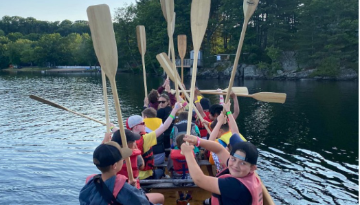 Du canotage au Centre Lake Joe! Des enfants et des jeunes sont assis dans le canoë Voyageur et tiennent leurs pagaies en l’air.