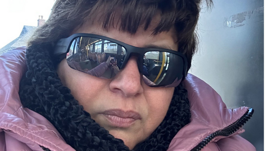 Marisa rides the bus. She’s wearing sunglasses, a pink winter coat and a black knitted scarf. The interior of a bus is reflected in her sunglasses.