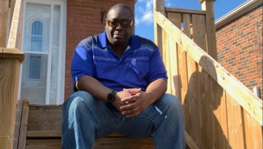 Martin sits on the porch steps outside his home. He’s wearing a blue polo t-shirt, jeans and sunglasses.