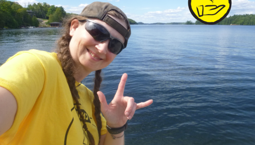Jessica Bailey pose sur le front de mer du lac Joe. Elle fait le signe en langue des signes américaine pour "Je t'aime". L'icône d'une main surmontée d'un cœur flottant apparaît dans le coin supérieur droit. 