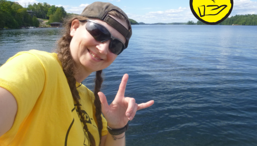 Jessica Bailey pose sur le front de mer du lac Joe. Elle fait le signe en langue des signes américaine pour "Je t'aime". L'icône d'une main surmontée d'un cœur flottant apparaît dans le coin supérieur droit.