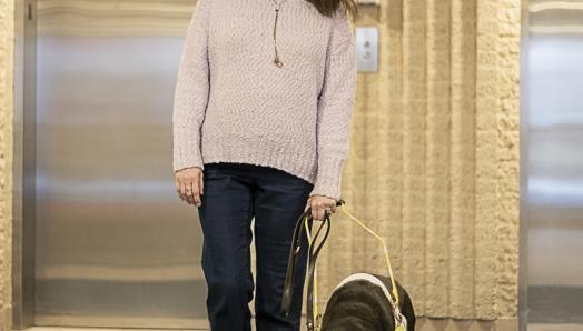 Karoline and her black guide dog, Raven, exit the lobby of a building. Behind them is a row of elevators.