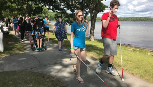 Two youth with white canes are walking along the lakeshore at CNIB Lake Joe. A group of additional youth are walking behind them.