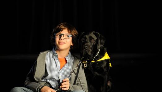 Gabriel sourit, assis à côté de Terry, son chien compagnon d'INCA, un Labrador retriever noir, sur le plateau de tournage du dernier message publicitaire de la campagne de collecte de fonds d'INCA.
