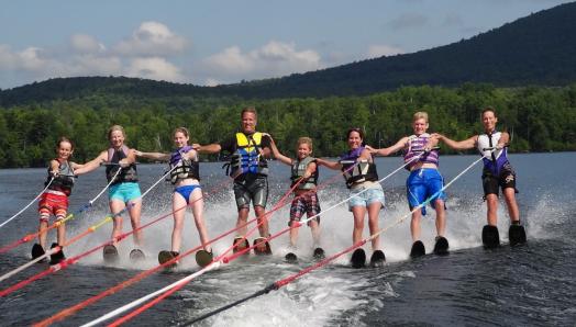 Une photo de 8 personnes — hommes, femmes, filles et garçons — faisant du ski nautique ensemble en ligne tout en ayant le bras sur la personne derrière eux.