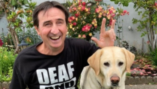 Eddy est assis devant un jardin dans une cour avec son chien-guide. Sa main gauche est au-dessus de la tête du chien et il signe « I love you » en langue des signes américaine. Le t-shirt noir qu’Eddy porte affiche le texte « Deaf and loud! » en gras. 
