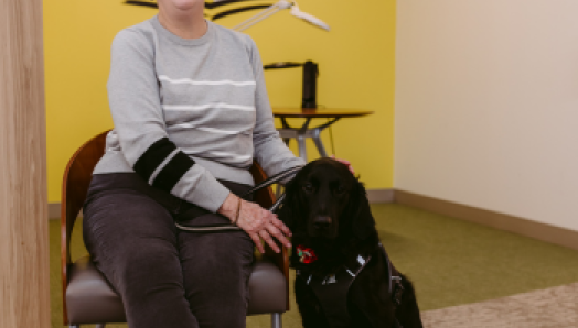 Dans un bureau d’INCA, Penny est assise sur une chaise. Son chien-guide, Honour, un croisement de labrador retriever noir et de golden retriever, est assis à ses pieds. Penny caresse la tête d’Honour et sourit de joie.