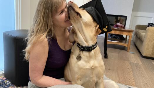 Laurie est assise sur le sol de son salon avec son chien-guide, Bridget, un croisement de labrador retriever jaune et de golden retriever. Bridget porte un chapeau de graduation et est assise sur les genoux de Laurie. Laurie presse affectueusement son visage contre le visage moelleux de Bridget.