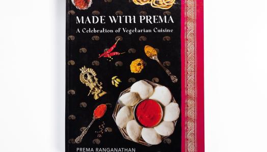 Une photo d’un livre de cuisine à couverture rigide intitulé Made with Prema apparaît sur un fond blanc uni. La couverture du livre de cuisine présente plusieurs plats sur un sari noir, or et magenta