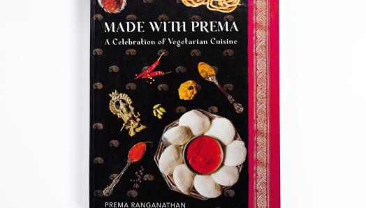 Une photo d’un livre de cuisine à couverture rigide intitulé  Made with Prema apparaît sur un fond blanc uni. La couverture du livre de cuisine présente plusieurs plats sur un sari noir, or et magenta