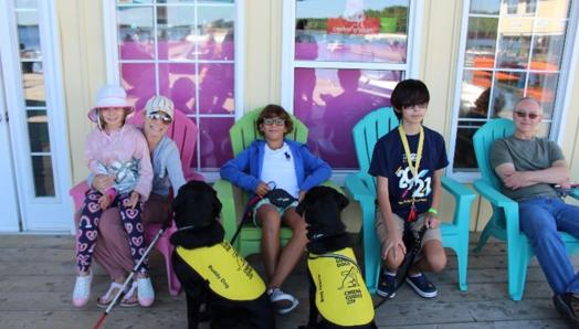 Gabriel, 12 ans, est assis dans une chaise Muskoka colorée, détendu et souriant, entouré d’autres familles et campeurs heureux du Centre Lake Joe d’INCA et de deux chiens compagnons d’INCA portant des gilets jaunes.