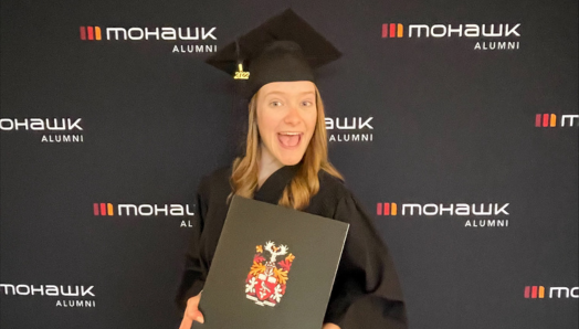 Graduation Day! Emilee, in her cap and gown, smiles and proudly displays her diploma. She is standing in front of a Mohawk College Alumni branded backdrop.