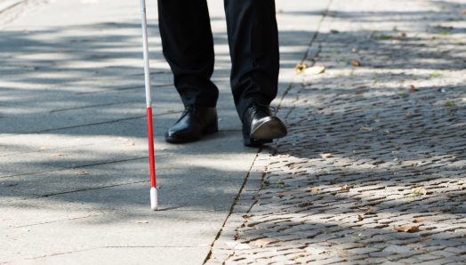 Une canne blanche balaie l’environnement sur un trottoir. À l'arrière-plan, des chaussures d'hommes traînent derrière le bout de la canne.