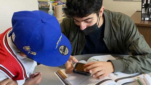 Dans une salle de classe, Aidan s’assoit avec un jeune élève et lui montre comment utiliser les fonctions d’agrandissement de son iPhone. Aidan tient son iPhone au-dessus d’un manuel scolaire. L’élève se penche attentivement pour assister à la démonstration.