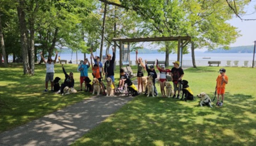 Une photo de groupe prise au bord de l’eau au Centre Lake Joe d’INCA. Le groupe est composé de participants avec des chiens compagnons. Ils lèvent les mains en l’air.