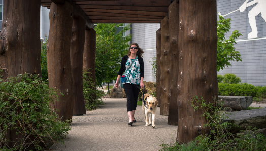 Larissa and their guide dog, a yellow lab named Piper, walk along an outdoor pathway at the Wayne Gretzky Sports Centre.