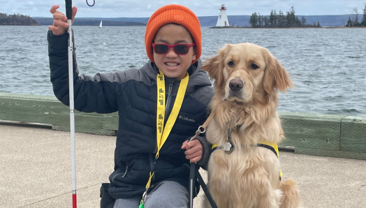 Emmett et Scout. Sur les rives d’un lac, Emmett est agenouillée sur le sol à côté de son chien compagnon Scout, un chien golden retriever à poil long. Emmett sourit et tient sa canne blanche dans sa main droite et la laisse de Scout dans sa main gauche.