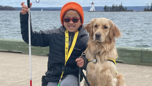 Emmett et Scout. Sur les rives d’un lac, Emmett est agenouillée sur le sol à côté de son chien compagnon Scout, un chien golden retriever à poil long. Emmett sourit et tient sa canne blanche dans sa main droite et la laisse de Scout dans sa main gauche. 