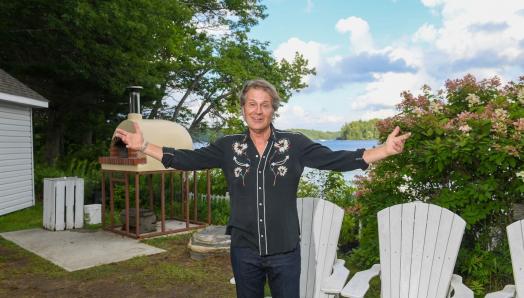 Jim Cuddy est debout, portant une chemise noire et des jeans, dans l’espace réservé au feu de camp, devant les chaises Muskoka blanches et le four à pizza en plein air. Il a les bras tendus et sourit.