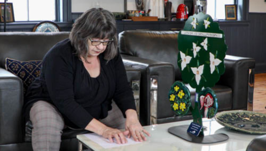 Robyn est assise sur une chaise revêtue de cuir et lit la description en braille d’une sculpture tactile que le canton d’Oro-Medonte lui a demandé de réaliser. La sculpture, posée sur la table, comporte des fleurs tactiles : des trilles, des boutons d’or et un cypripède.