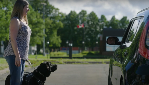 Une femme, debout sur le trottoir avec son chien-guide, parle au chauffeur de taxi qui s’est arrêté à côté d’elle.