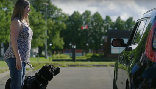 Une femme, debout sur le trottoir avec son chien-guide, parle au chauffeur de taxi qui s’est arrêté à côté d’elle. 