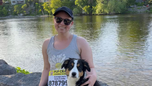 Keely et son border collie chiot sont assis sur un rocher au bord d'un lac. Keely porte un dossard de la course à pattes d'INCA et passe le bras autour de son chiot.