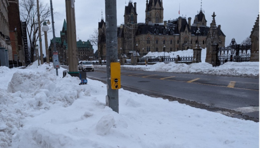An APS button fixed to a pole at a crosswalk in Ottawa, with Parliament buildings in the background. The pole is surrounded by a large snowbank that reaches almost as high as the button and extends approximately a metre outward, making it impossible for pedestrians to reach it.