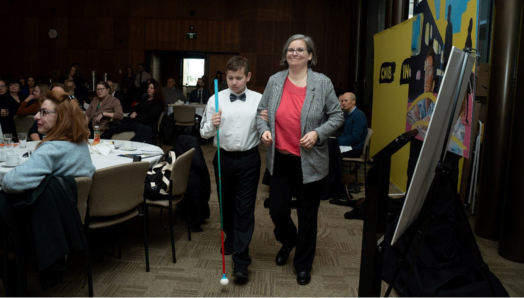 Dawn Pickering sourit en se dirigeant avec son fils, Ollie, vers la scène pour le panel des familles lors du lancement parlementaire de la Charte des droits des enfants d’INCA. On voit les participants devant une grande bannière d'INCA à l'arrière-plan. Ollie porte une chemise blanche avec un nœud papillon noir et tient sa canne blanche.