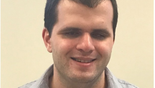 A headshot of Rilind Dragoshi smiling. He has short dark hair and wears a collared shirt in front of a white background