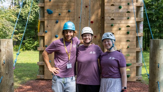 Three young CNIB Lake Joe staff members stand in front of the new climbing tower at Lake Joe. They are wearing helmets and CNIB Lake Joe staff shirts.