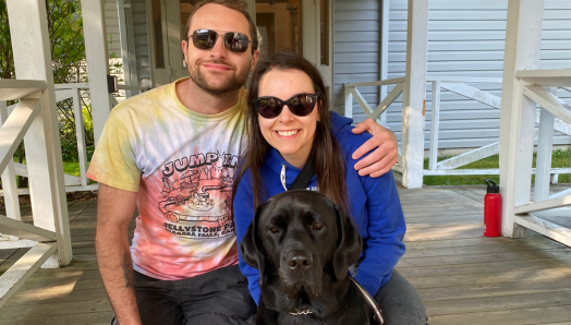 At CNIB Lake Joe, Micheal and Danica kneel on the boardwalk with a black Labrador Retriever. Picture taken during the Orientation & Mobility/Guide Dog Immersion program at CNIB Lake Joe.